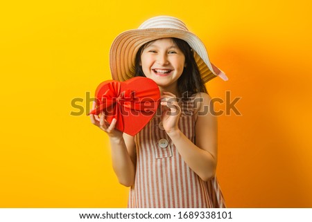 Portrait of a little pretty girl and summer striped dress and hat holding a red heart-shaped gift box. Cheerful and happy child posing isolated on yellow studio background.