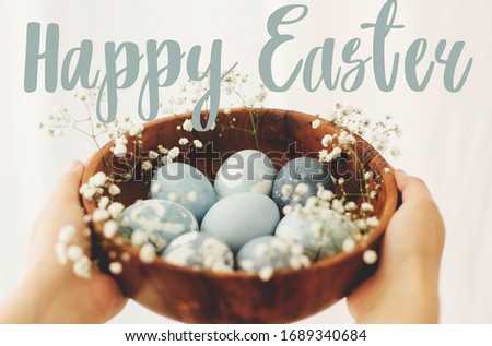 Happy Easter text. Easter greetings lettering. Hands holding wooden bowl with modern blue Easter eggs in spring flowers. Greeting card
