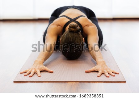 Yogi woman in balasana over bright white background. Hands of lady extended on pink mat and body resting on legs at studio. Healthy lifestyle, stretching exercise, wellness, yin yoga concepts Royalty-Free Stock Photo #1689358351