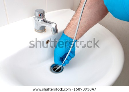 Plumbing issues, occupation in sanitation and handyman contractor concept with plumber repairing drain with plumbers snake (steel spiral that twists through pipes to collect dirt) in residential sink Royalty-Free Stock Photo #1689393847