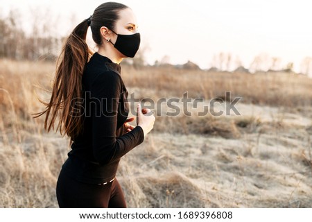 Sport during quarantine, self-isolation in the countryside. A young athletic woman is jogging on a dirt road in the meadow. He is wearing a black medical mask and headphones. Photo in motion