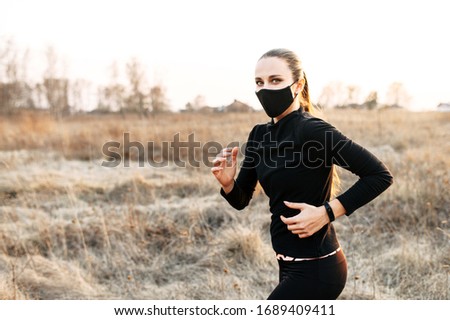 Sport during quarantine, self-isolation in the countryside. A young athletic woman is jogging on a dirt road in the meadow. He is wearing a black medical mask and headphones. Photo in motion