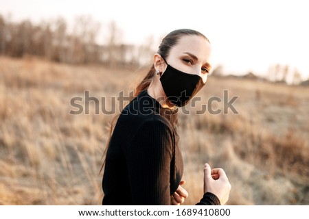 The concept of sports training during quarantine. Young woman in a black sports outfit and in a black medical mask outdoors, Photo at sunset in countryside