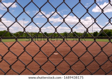 Look out at a baseball field through the diamond patterned grid of a chain link fence from behind home plate on a beautiful spring day with partly cloudy blue skies.