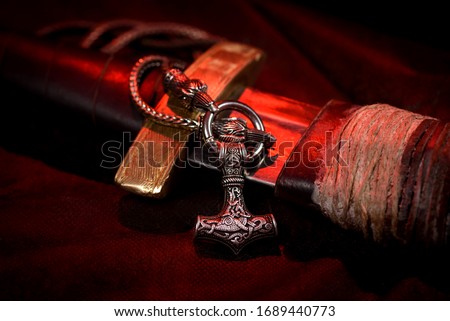 Viking battle sword made of steel with a leather handle with a Thor's silver Hammer pendant on a red black background. Handmade. Mjolnir. Viking's symbol Royalty-Free Stock Photo #1689440773