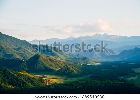 Wonderful mountain land with forest and small houses in sunlight. Scenic view to vast expanses of rolling hills and great mountain ridge on horizon. Immense distances of wavy relief in evening light. Royalty-Free Stock Photo #1689510580