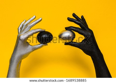 Silver and black eggs holding in woman’s chrome and black matte hands. Isolated on yellow background .Concept ideas.