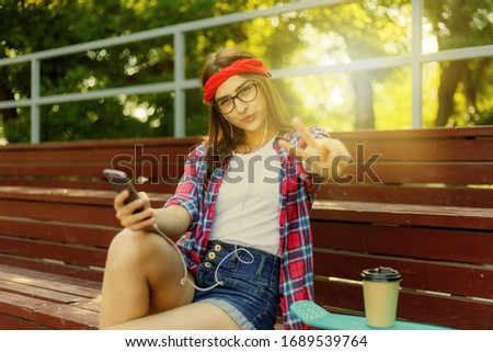 Young woman skater dressed in stylish clothes sits in the stands in a skatepark holds a smartphone in her hands and listens to music in earphones on a bright sunny day