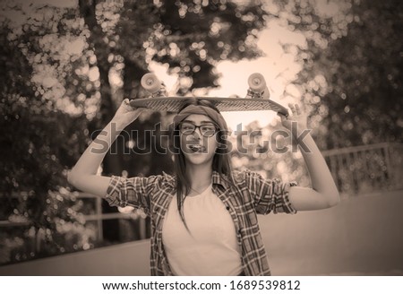 Funny young hipster woman dressed in stylish clothes holds skateboard in her hand in skatepark at bright sunny day. Summer lifestyle image of trendy pretty young girl 
