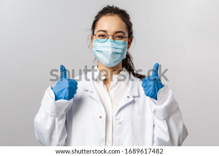 Doctors, infectionist, research and covid19 concept. Satisfied young asian female doctor receive good results on studying virus, discover vaccine, show thumb-up, wear face mask and gloves Royalty-Free Stock Photo #1689617482