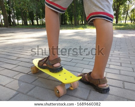 Feet of a little boy in a brown sandals on a yellow children's skateboard in the park. Close view. Summer activity concept.