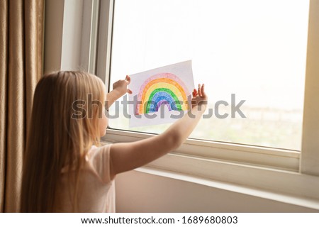Kid painting rainbow during Covid-19 quarantine at home. Girl near window. Stay at home Social media campaign for coronavirus prevention, let's all be well, hope during coronavirus pandemic concept Royalty-Free Stock Photo #1689680803