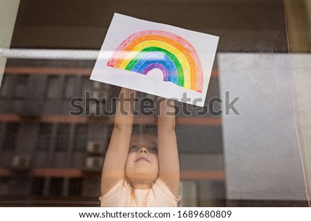 Kid painting rainbow during Covid-19 quarantine at home. Girl near window. Stay at home Social media campaign for coronavirus prevention, let's all be well, hope during coronavirus pandemic concept Royalty-Free Stock Photo #1689680809
