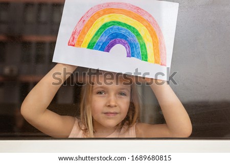 Kid painting rainbow during Covid-19 quarantine at home. Girl near window. Stay at home Social media campaign for coronavirus prevention, let's all be well, hope during coronavirus pandemic concept