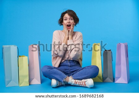 Indoor photo of amazed young dark haired curly lady holding raised palms on her cheeks while looking at camera, making too much purchases while posing over blue background