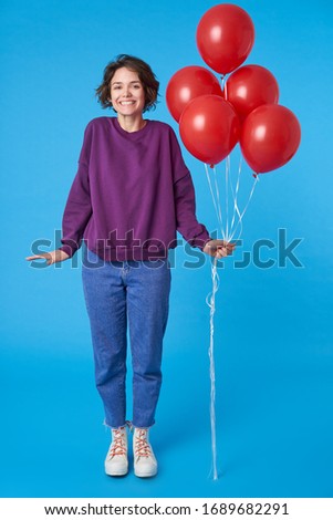 Happy young beautiful dark haired woman holding bunch of red air balloons and looking cheerfully at camera, celebrating something while posing over blue background