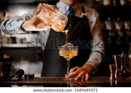 barman gently pours finished cocktail from glass shaker into glass. Body of bartender in black apron on background. Royalty-Free Stock Photo #1689689371