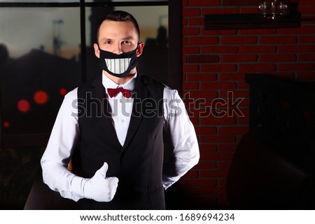 A young waiter with a cheerful protective mask on his face. Health protection in the restaurant. The problem of spreading the virus. COVID-19