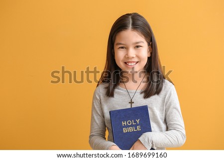 Cute little Asian girl with Bible on color background