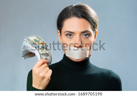 a young lady in black holds in her hand a lot of cash American dollars with banknotes of one hundred dollars, close-up portrait with her mouth closed with a white ribbon. Royalty-Free Stock Photo #1689705190