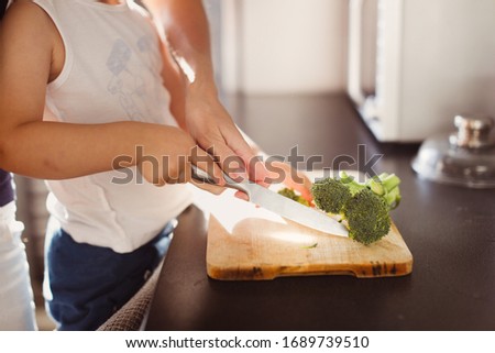 Mom with the baby in her arms help the boy son chop the broccoli on his own. Proper healthy nutrition and help to mom, the child cuts himself with knife