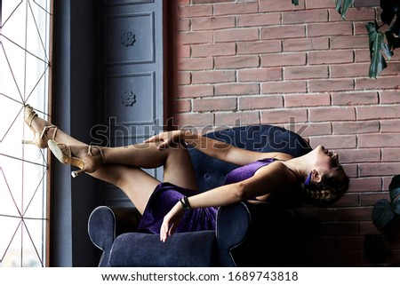 Young brunette woman, wearing dark violet top and skirt, sitting relaxing lying in dark blue navy armchair in loft living room with brick wall and green plant. Salsa dancer female photo shoot.