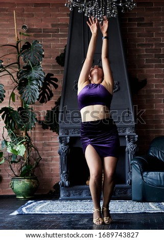 Young brunette woman, wearing dark violet top and skirt, doing dance posing in dark loft living room with brick wall, old black fireplace and green plant. Salsa dancer female photo shoot.