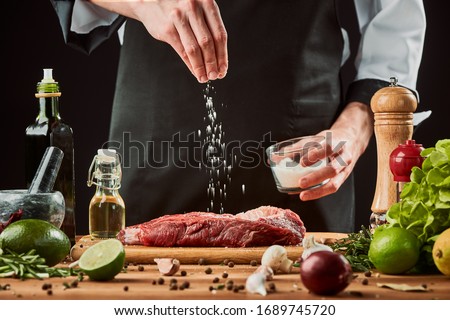 Man sprinkles salt over meat. Chef cooking a beef steak, seasoning with pepper, lime, olive oil, garlic and onion. Royalty-Free Stock Photo #1689745720