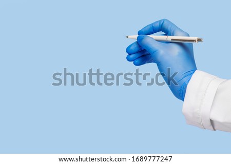 A female hand in a medical glove and in a white coat holds a pen and writes in the air on a blue background.