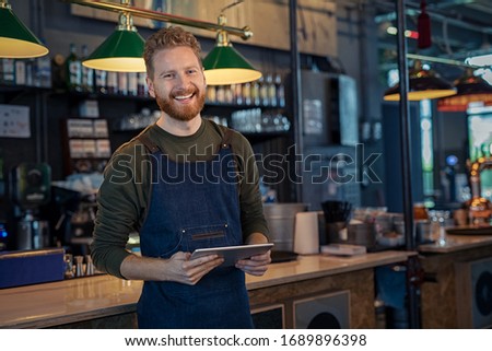 Successful small business owner using digital tablet and looking at camera. Happy smiling waiter ready to take order. Portrait of young entrepreneur of coffee shop standing at counter with copy space. Royalty-Free Stock Photo #1689896398
