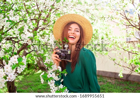 Playful cute girl making funny face and making photos with vintage camera , posing in spring park on blooming trees background.Carefree girl in stylish straw hat and green dress enjoying sunny day.