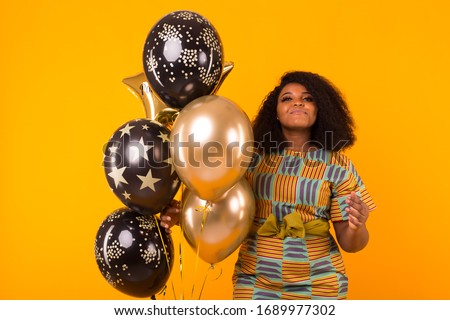Holidays, birthday party and fun concept - Portrait of smiling young African-American young woman looking sweet on yellow background holding balloons.