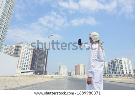 Middle east man wearing traditional white suit with head scarf taking picture of buildings with smartphone on city street over sky background, Bahrain.