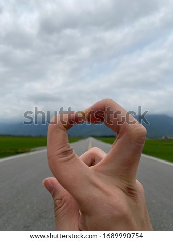 Bending fingers with a hart shape with a beautiful country road and mountain background. 