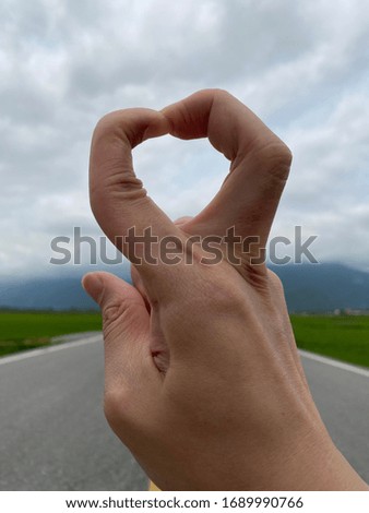 Bending fingers with a hart shape with a beautiful country road and mountain background. 