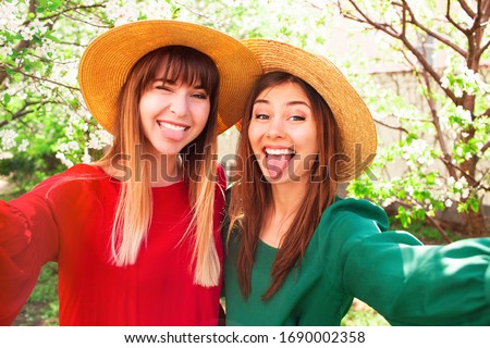 Close up lifestyle portrait of two pretty girls, best friends making self portrait by camera at blossom tree. Wearing similar summer straw hats. Sunny spring day, showing tongue, crazy emotions.