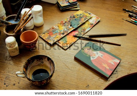 Cup covered with paint, with a wood stain substance. Reproduction of the icon of St. Nicholas. The process of the artist's work. Brushes and paints on the background in studio.