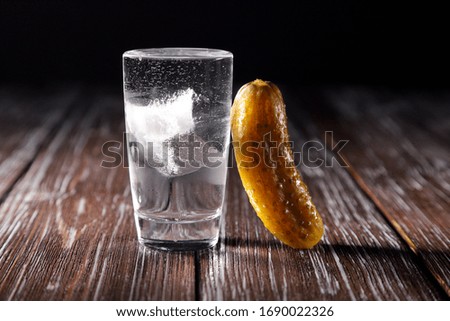 A full stack of cold vodka with ice and pickled cucumber, on a wooden table and black background.