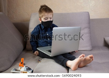 little boy sitting on a sofa in a black medical mask and looking at a laptop