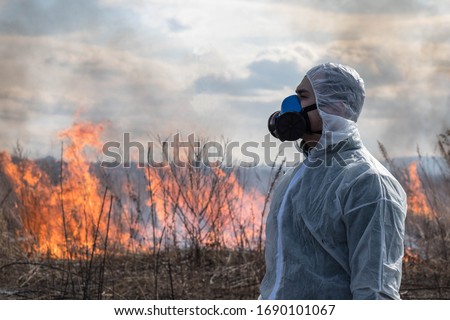 Portrait of a man in a white chemical protective suit and a respirator against the background of a forest fire. Concept of environmental, chemical, epidemic and radiation problems. Royalty-Free Stock Photo #1690101067