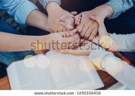 Close up of  people group holding hand and pray together over a blurred holy bible on wooden table, christian fellowship  or praying meeting in home concept with copy space