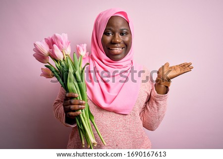 Young african american plus size woman wearing muslim hijab holding bouquet of pink tulips smiling cheerful presenting and pointing with palm of hand looking at the camera.