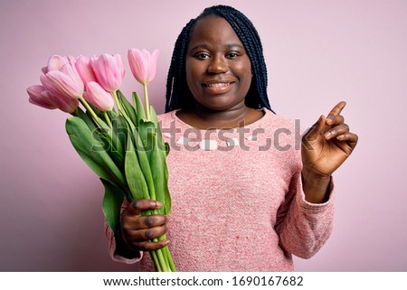 Young african american plus size woman with braids holding bouquet of pink tulips flower cheerful with a smile on face pointing with hand and finger up to the side with happy and natural expression