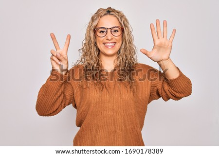 Young beautiful blonde woman wearing casual sweater and glasses over white background showing and pointing up with fingers number seven while smiling confident and happy.