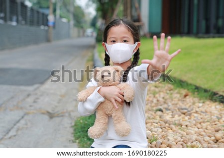 Asian little girl show palm sign wearing mask to protect corona virus and air pollution dust with hugging teddy bear doll sitting outdoor. Stop Coronavirus covid-19 and pollution protection concept.