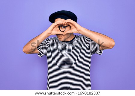 Handsome man with blue eyes wearing striped t-shirt and french beret over purple background Doing heart shape with hand and fingers smiling looking through sign