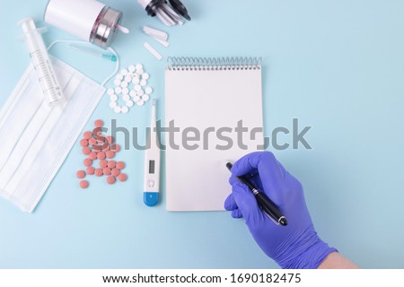 Evidence based medicine concept.Treatment protocols. Doctor's hand in blue medical glove write a prescription. Blue desk with pills, thermometr, medicine mask, syringe. Mock up blank empty notepad.  Royalty-Free Stock Photo #1690182475