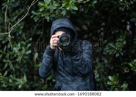A male photographer takes a photo with a digital camera while wearing a hoodie and hiding in the bushes in a forest.