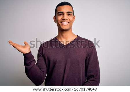 Young handsome african american man wearing casual sweater over white background smiling cheerful presenting and pointing with palm of hand looking at the camera.