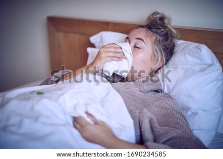 Woman with flu in bed as she blows her nose and looks at a thermometer to check her temperature 